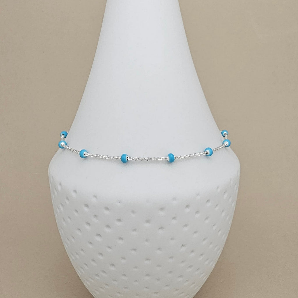 A delicate silver chain anklet with small light blue beads, displayed on a white textured cone jewelry stand against a beige background.