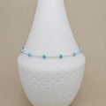A delicate silver chain anklet with small light blue beads, displayed on a white textured cone jewelry stand against a beige background.