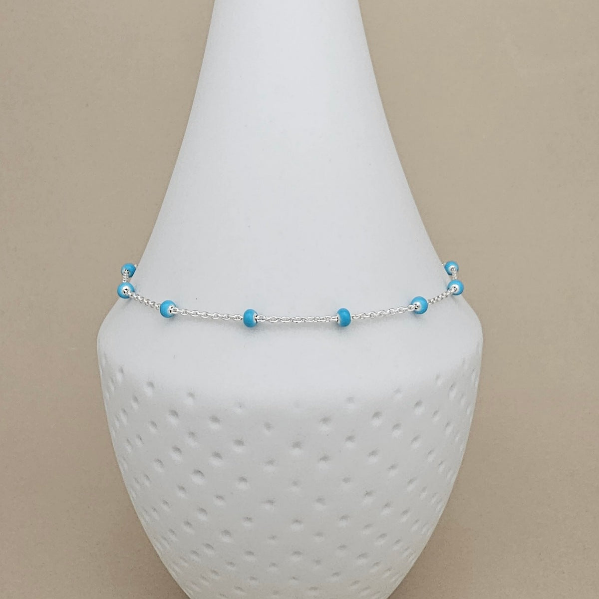A delicate silver chain anklet with small light blue beads, displayed on a white textured cone jewelry stand against a beige background.
