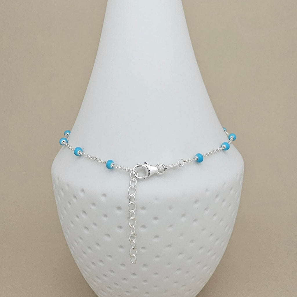 A delicate silver chain anklet with small turquoise blue beads, displayed on a white textured cone stand against a neutral beige background.
