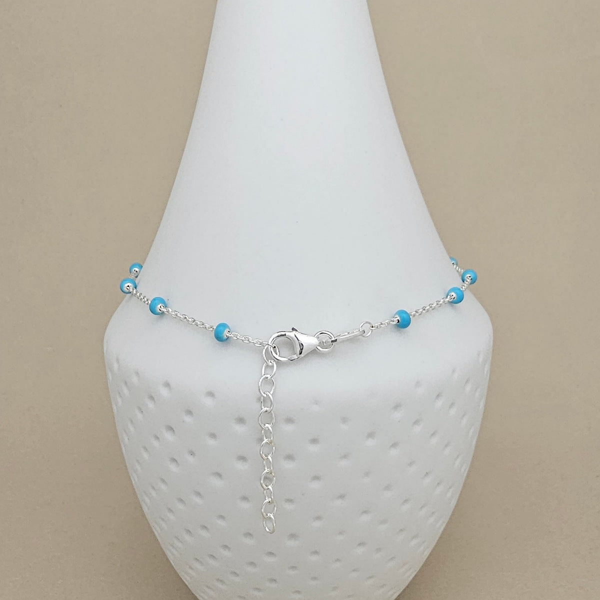 A delicate silver chain anklet with small turquoise blue beads, displayed on a white textured cone stand against a neutral beige background.