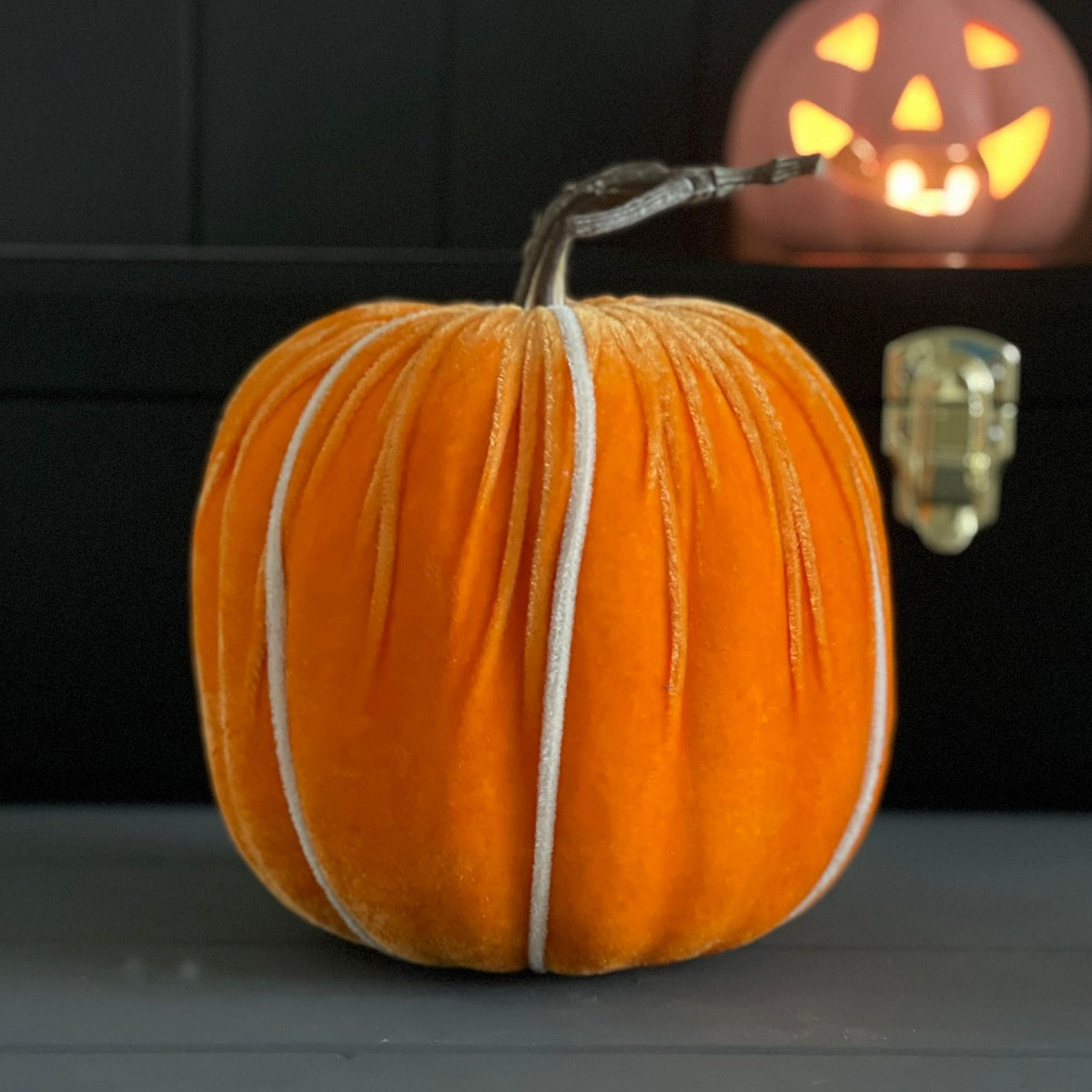 An orange velvet pumpkin with white faux stripes and a brown stem, displayed on a dark surface with a glowing jack-o'-lantern in the background.