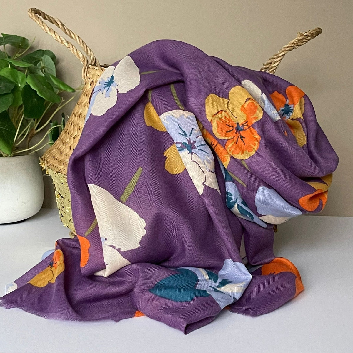 A lightweight dusky purple scarf with a vibrant viola floral print in yellow, orange, cream and white, draped over a woven straw basket with a green plant in the background.