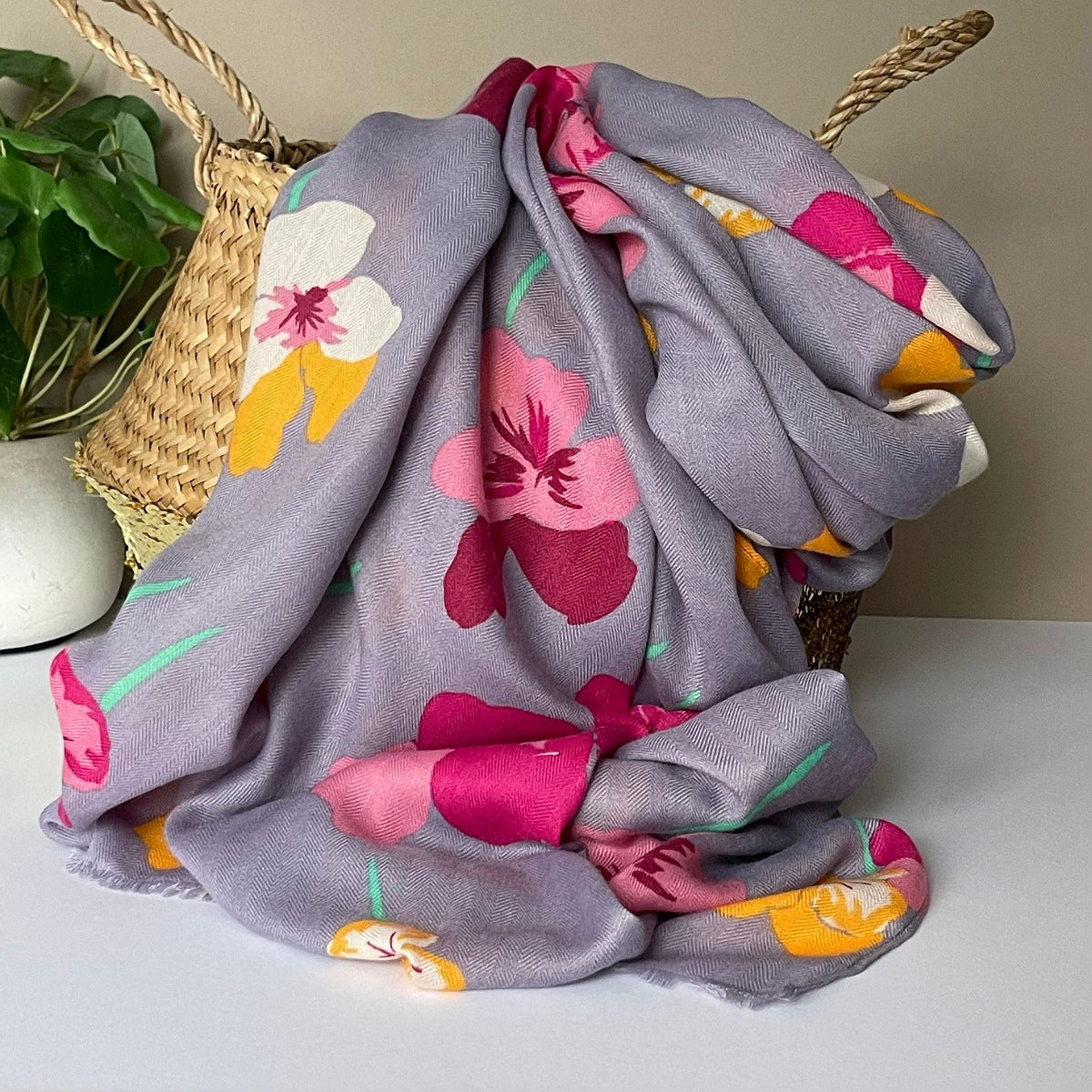A lightweight grey scarf with a vibrant viola floral print in pink, yellow, and white, draped over a woven straw basket with a green plant in the background.