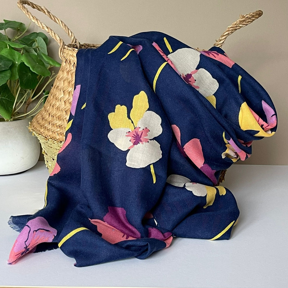 A navy blue scarf with a viola floral print in yellow, pink, and white, draped over a woven straw basket next to a potted green plant.
