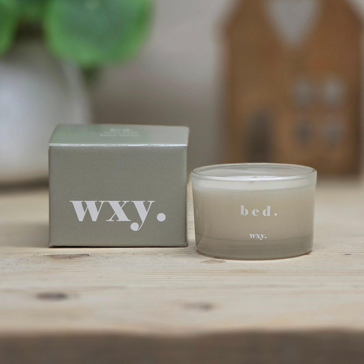 A pale grey candle in a frosted glass jar next to its grey packaging box, placed on a light wooden surface with a softly blurred background.