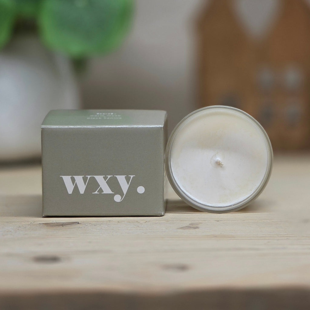 A white wax candle in a clear glass jar sits beside its matte grey-sage green box, both printed with the brand name 'wxy.', on a light wooden tabletop with a softly blurred background.