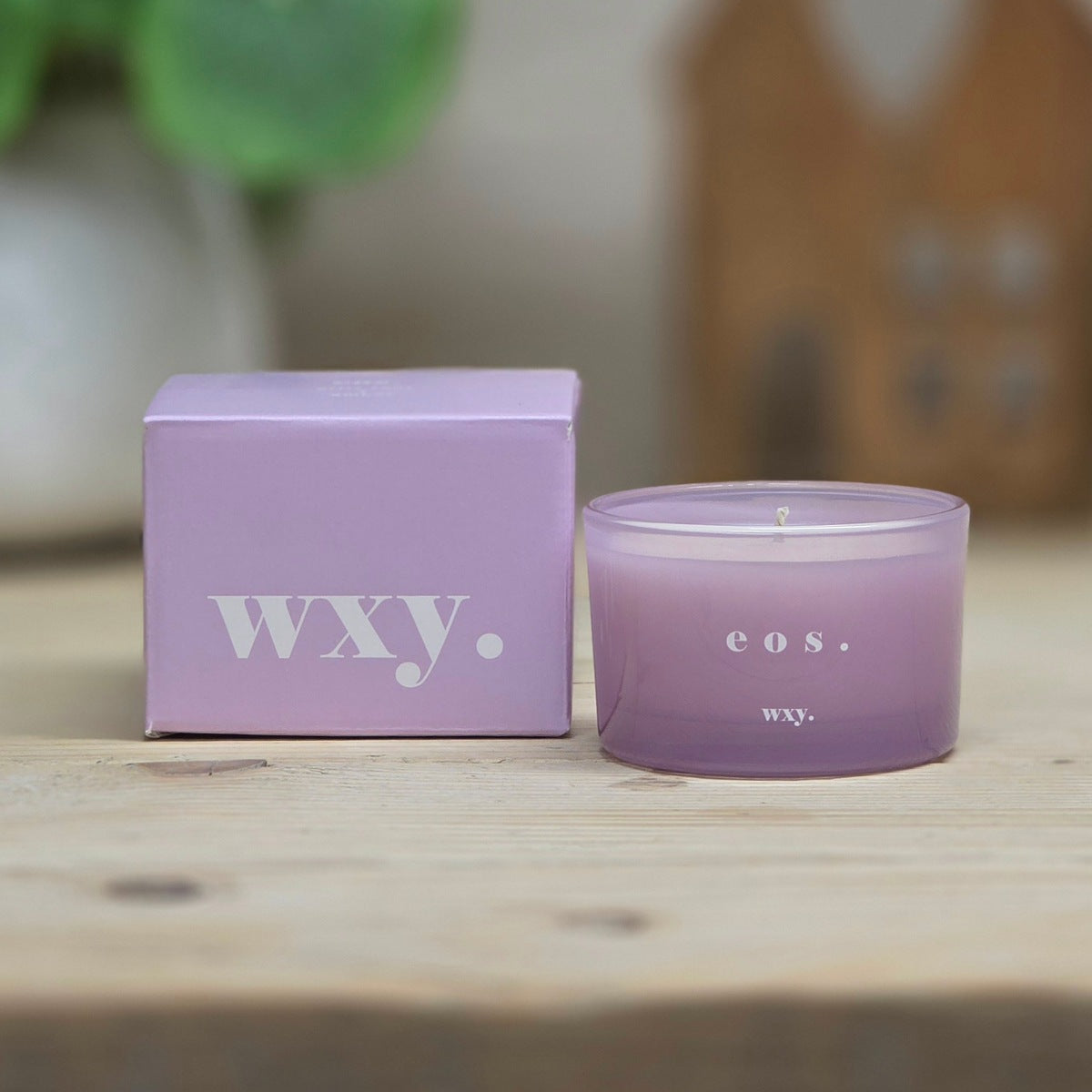 A lavender purple coloured candle in a clear glass jar sits next to its matching light purple packaging box on a light wooden surface. The box and jar are both printed with the brand name 'wxy.