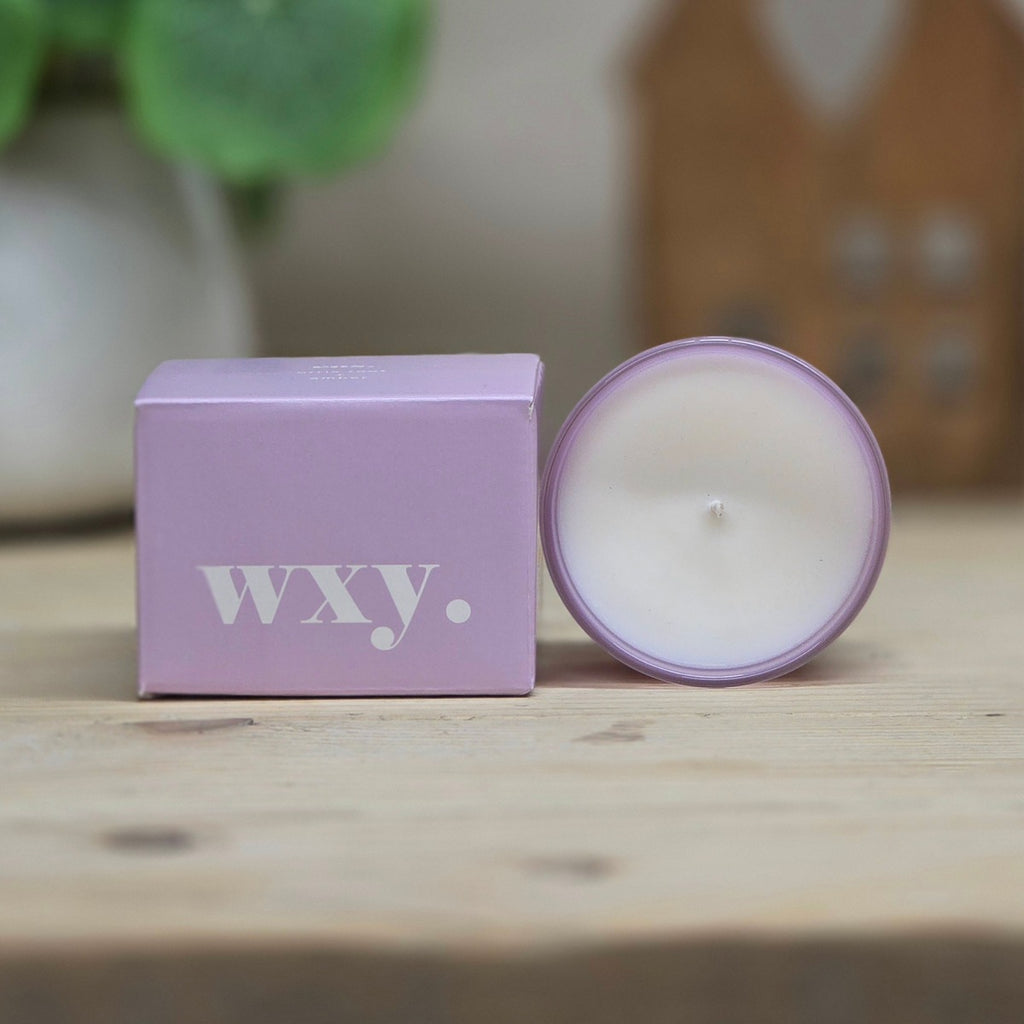 A lavender purple coloured candle in a round glass jar next to its matching purple box, both featuring the white 'wxy.' logo, set on a light wooden surface with a blurred background.