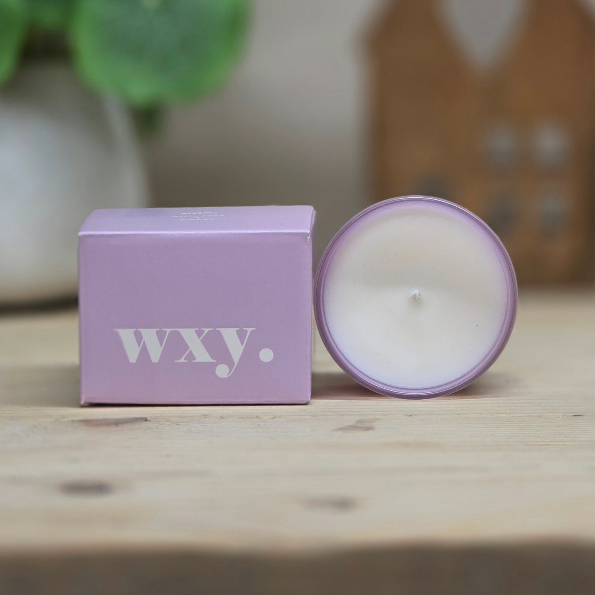 A lavender purple coloured candle in a round glass jar next to its matching purple box, both featuring the white 'wxy.' logo, set on a light wooden surface with a blurred background.