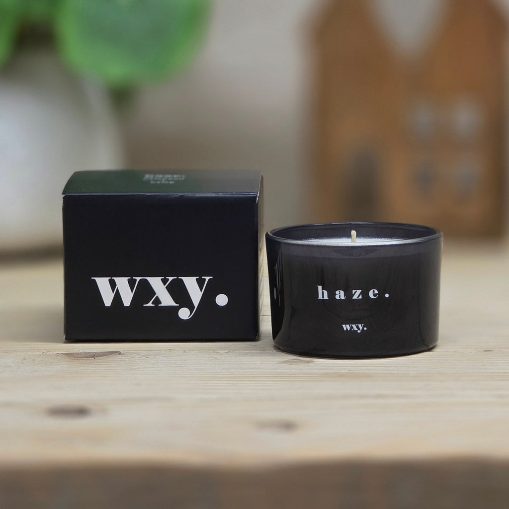 A black wax candle in a matte black jar next to its matching black packaging box, both printed with the white text 'wxy.' and 'haze.