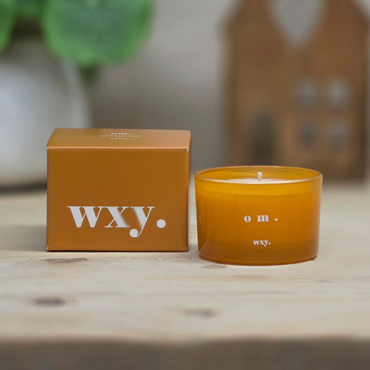 An amber glass jar candle next to its matching brown cardboard box, both featuring the white 'wxy.' logo, displayed on a light wooden surface with a softly blurred background.
