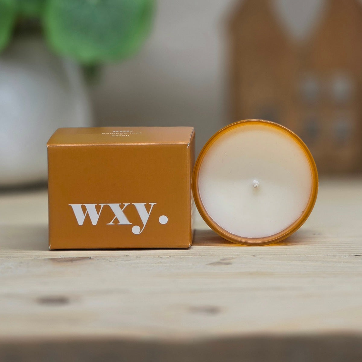 A WXY candle shown with its matching brown cardboard box on a light wooden surface. The candle is in a round amber-colored glass container, with the white wax visible and the wick centered.