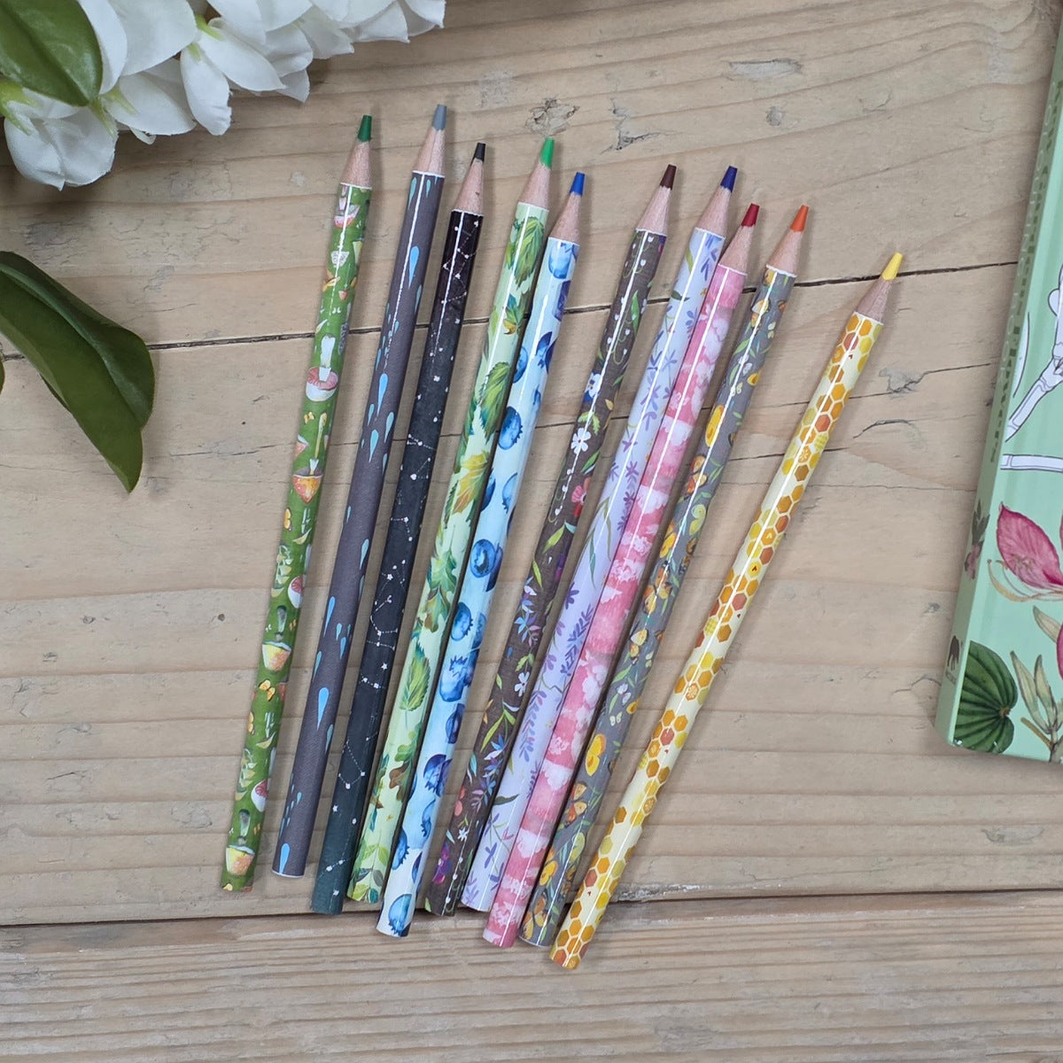 Set of colorful patterned pencils on a wooden surface with a box and flowers in the background.