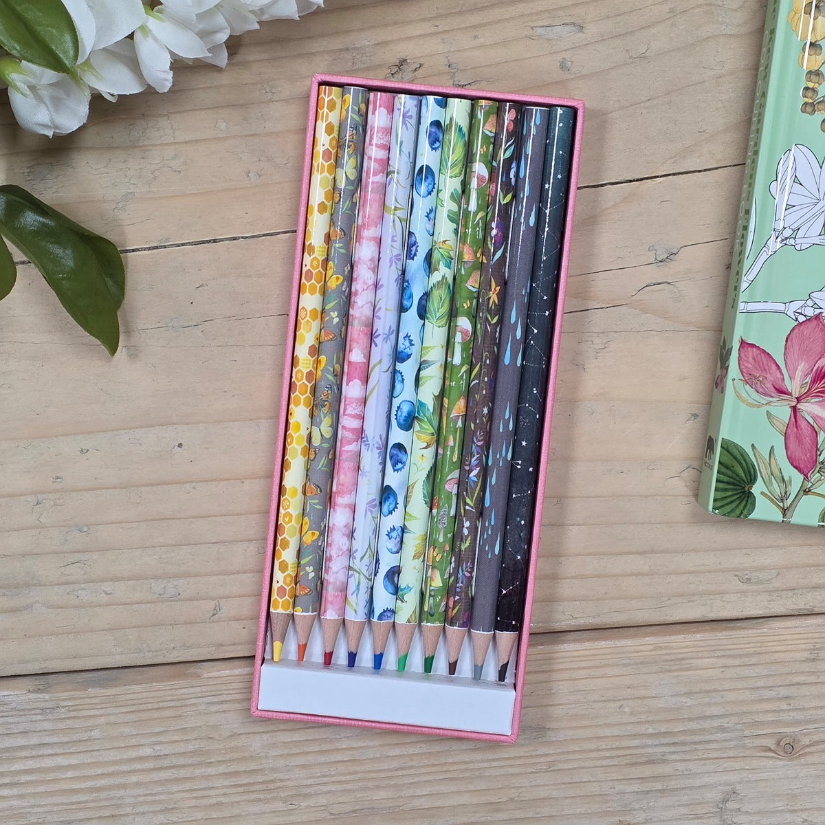 A pink rectangular pencil case holding eight patterned colored pencils, placed on a light wood surface with white flowers and part of another floral item visible at the edges.