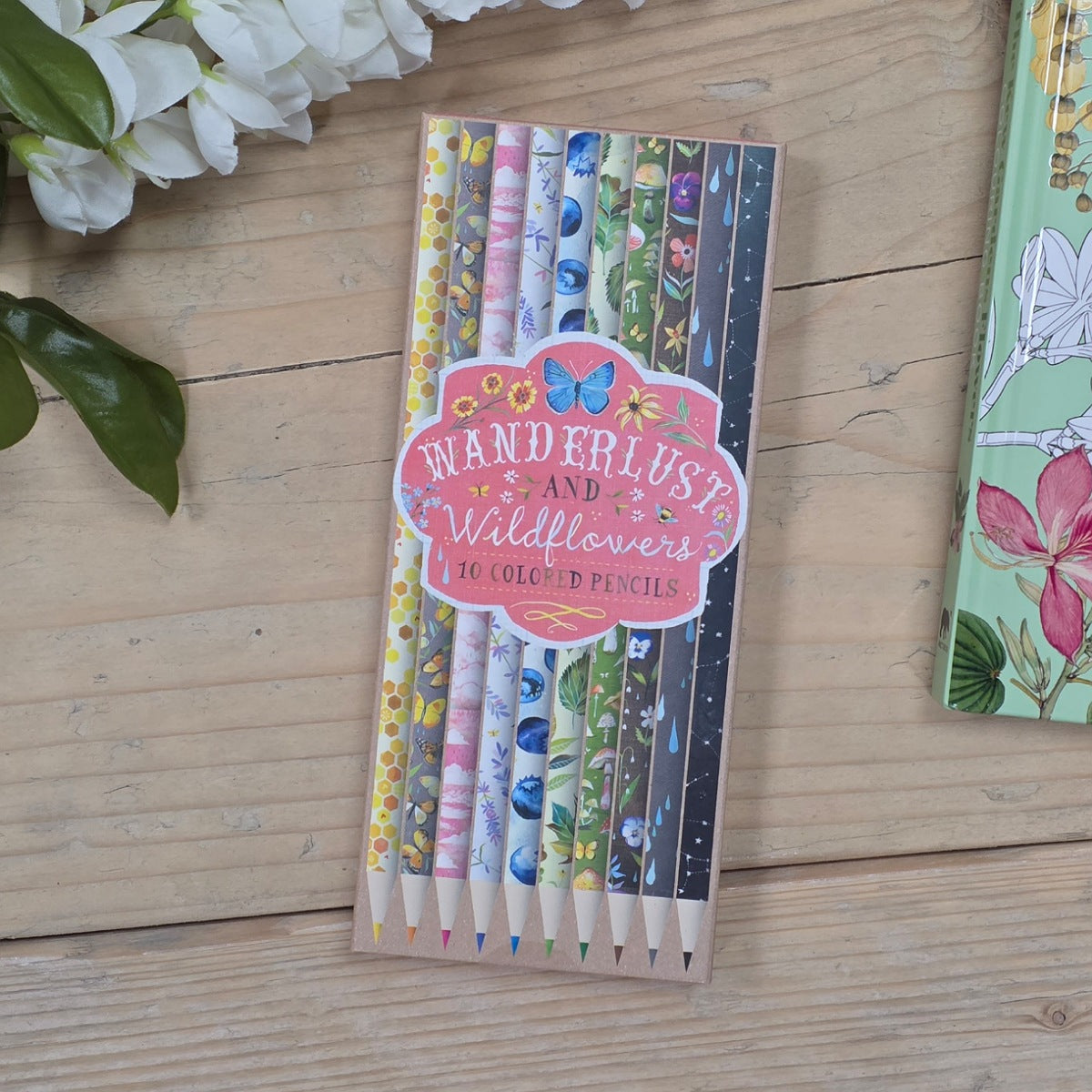 A set of 10 colored pencils in a clear rectangular box with a pink label reading 'Wanderlust and Wildflowers 10 Coloured Pencils,' placed on a light wood surface with white flowers nearby.