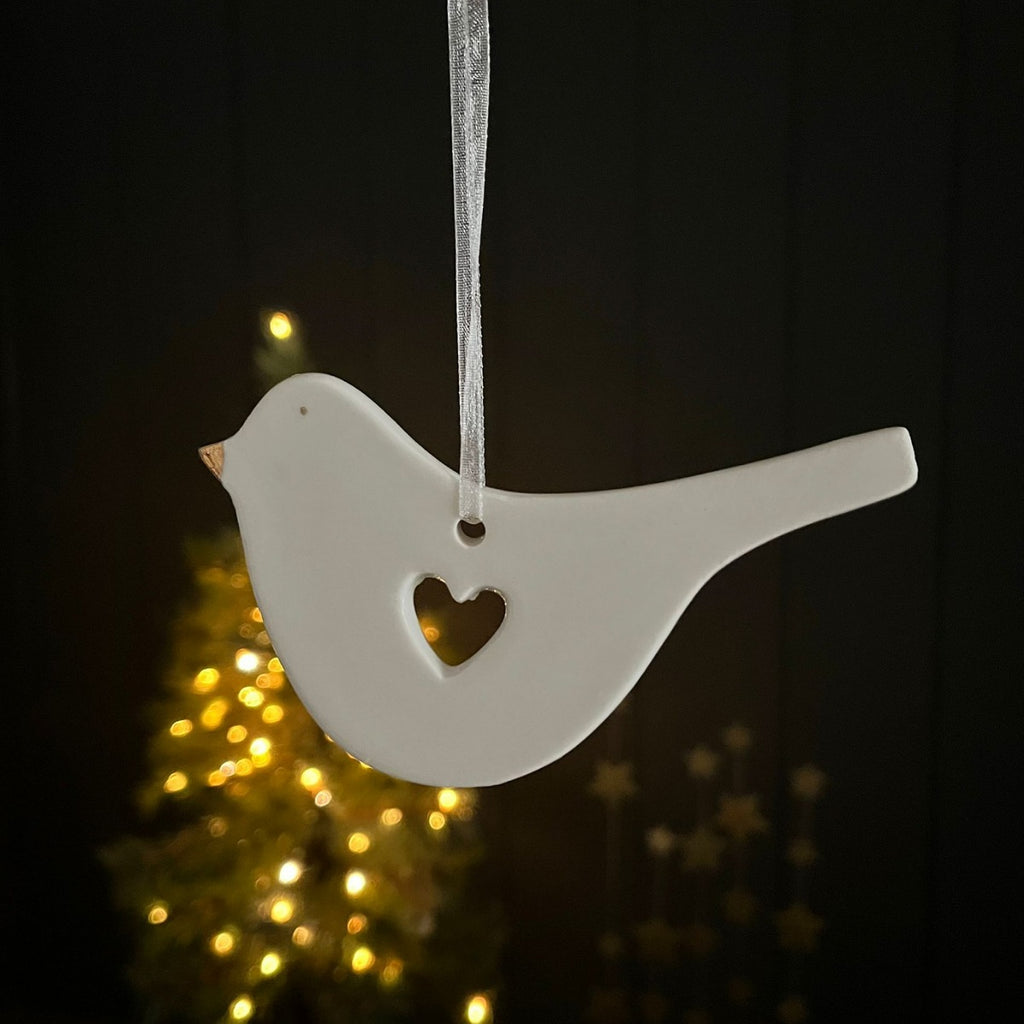 White ceramic bird ornament with a heart cutout in the middle hanging against a dark background with blurred lights.