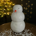 A glossy white ceramic snowman figure with a small orange carrot nose and black dot eyes, standing on a wooden surface with white decorative beads scattered around it. A lit Christmas tree and gold star decorations are blurred in the background.