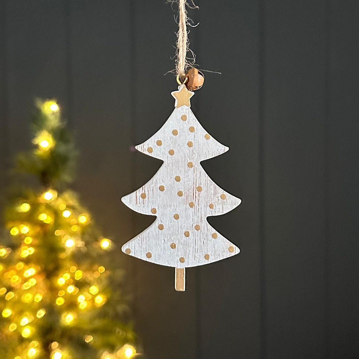 Wooden hanging tree decoration in white with gold dots, a star to the top with a gold bell.  Hanging with a dark background a blurred out Christmas tree.