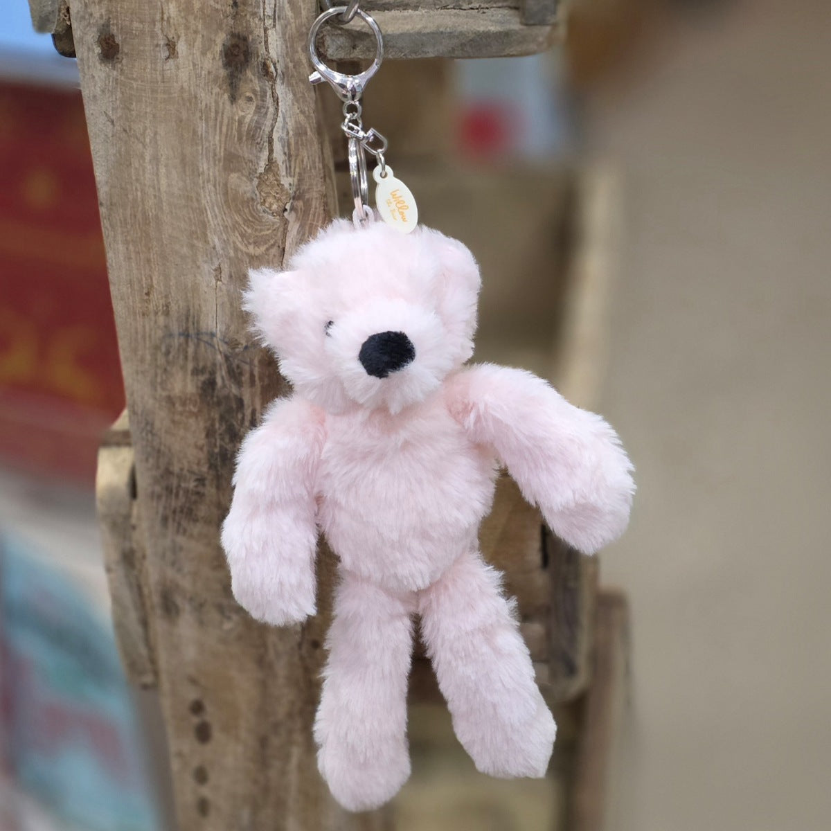 A small plush teddy bear keychain in light pink faux fur, hanging from a silver metal clasp on a rustic wooden post.