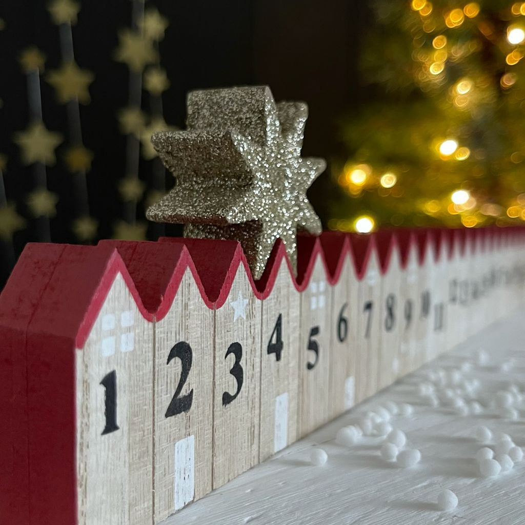 Wooden advent calendar with numbered houses and red roofs with a moveable gold star on top, Christmas tree in the background.