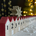 Wooden advent calendar with numbered houses and red roofs with a moveable gold star on top, Christmas tree in the background.