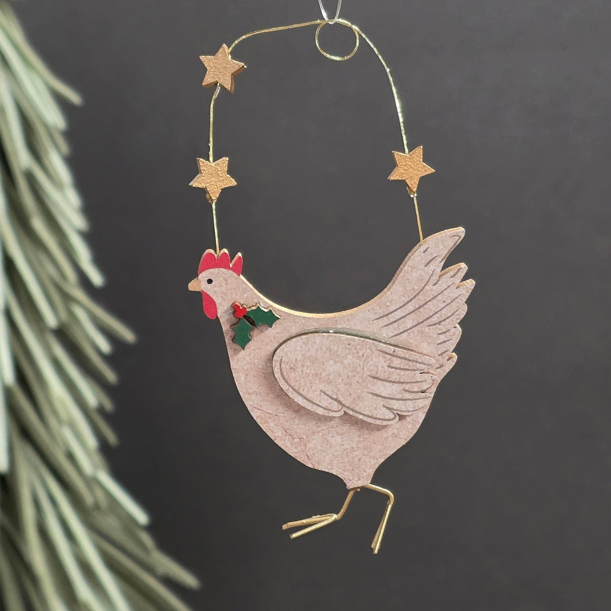 A wooden chicken hanging ornament with a gold wire hanger, star accents, and a small red and green holly detail, shown against a dark background with blurred pine branches on the left.
