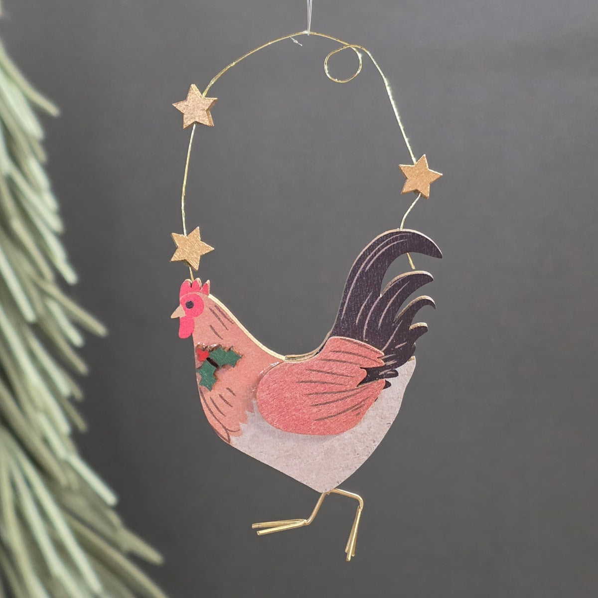 A wooden rooster Christmas ornament hanging from a gold wire hoop decorated with gold stars. The rooster features a pink and peach body, a white belly, a black tail, and a small green holly accent on its chest, set against a dark grey background with blurred pine needles on the left.