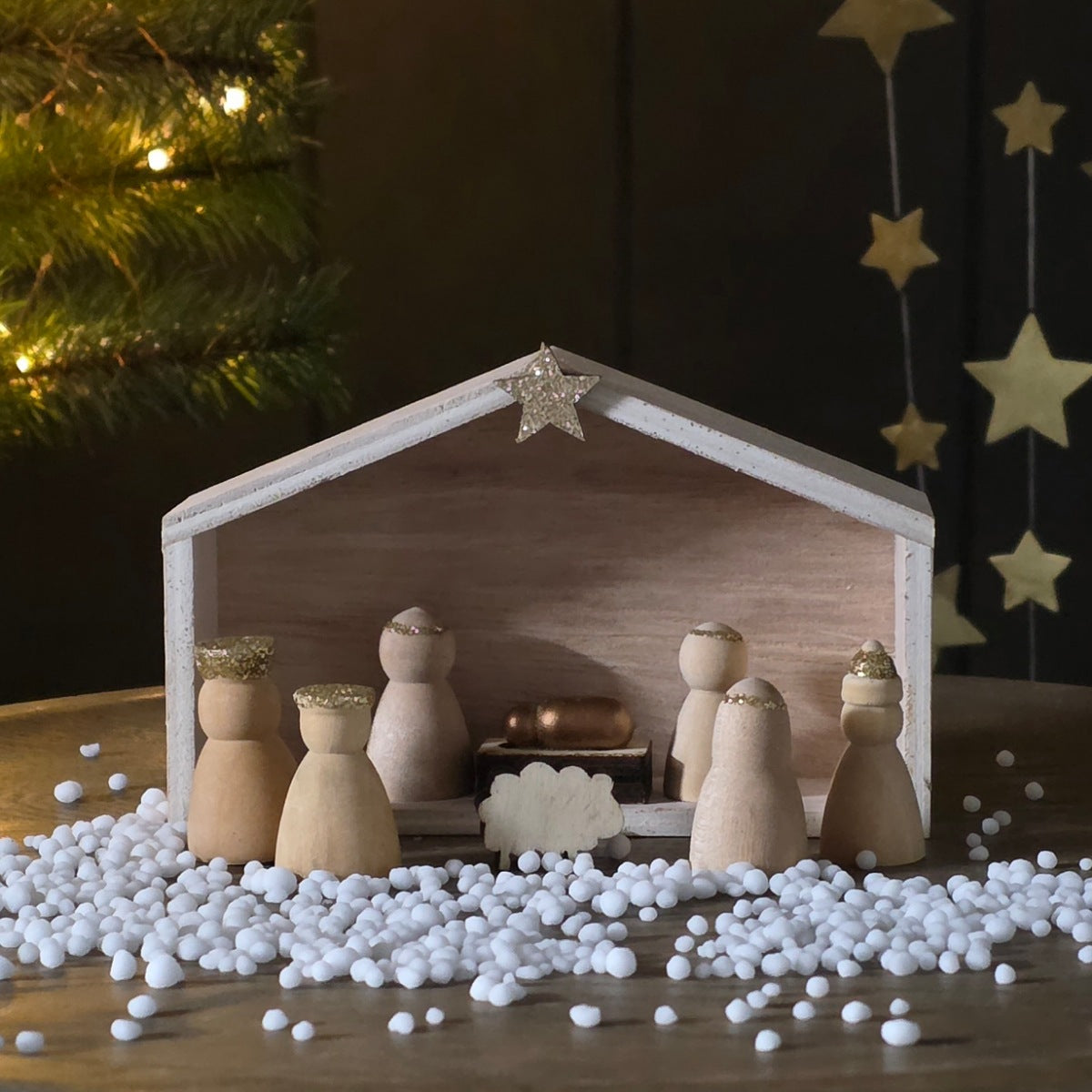 A wooden nativity scene set on a tabletop with small white decorative pebbles in front. The scene includes a light wood stable frame, several small wooden figures, a small manger or cradle piece, and a gold star ornament on the roof.