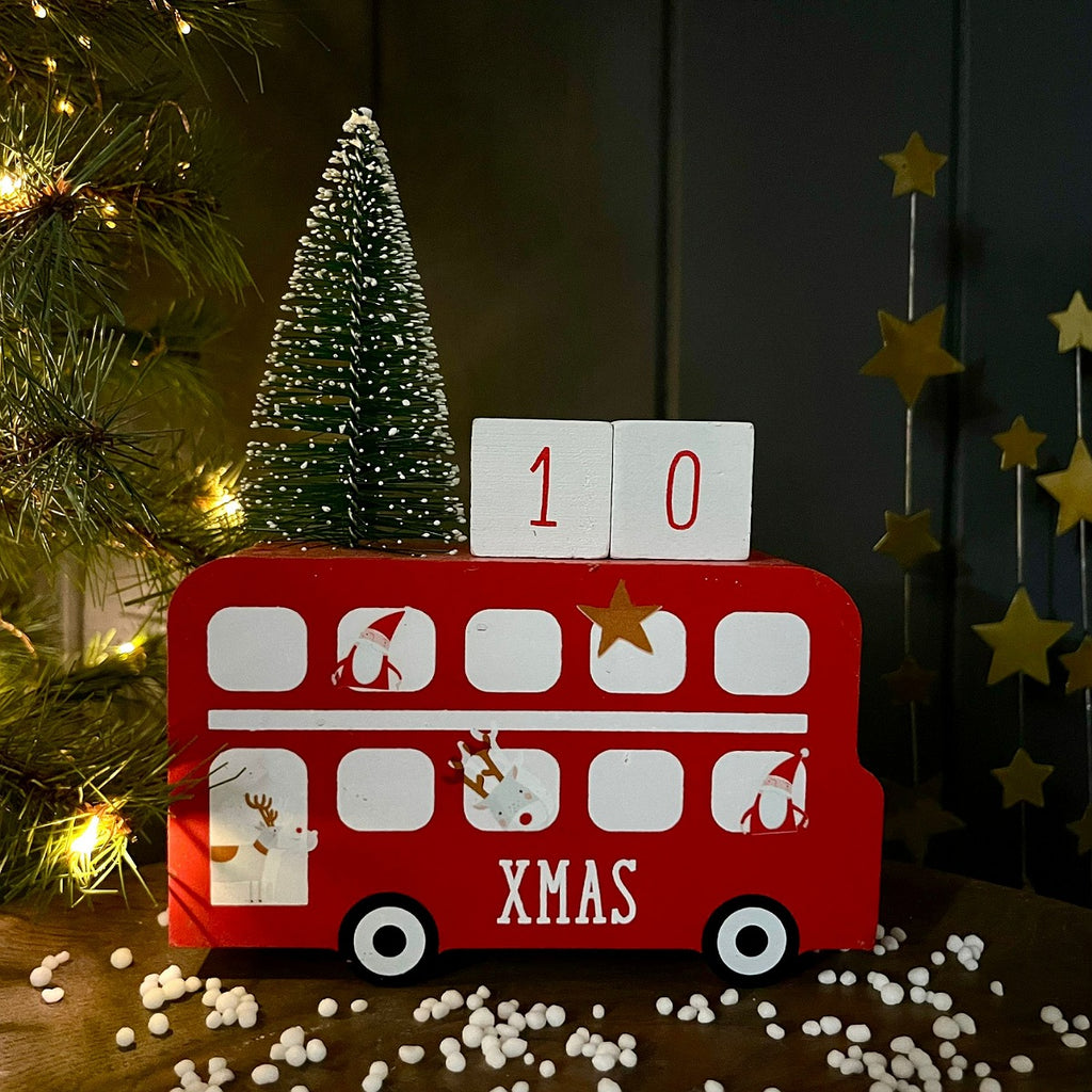 A red double-decker Christmas bus-shaped decorative calander with white windows, white reindeer and Santa illustrations, the word 'XMAS' on the front, and two white blocks showing '10' on top. It is styled on a tabletop with small white stones, a miniature Christmas tree, warm string lights, and hanging gold stars against a dark panelled background.