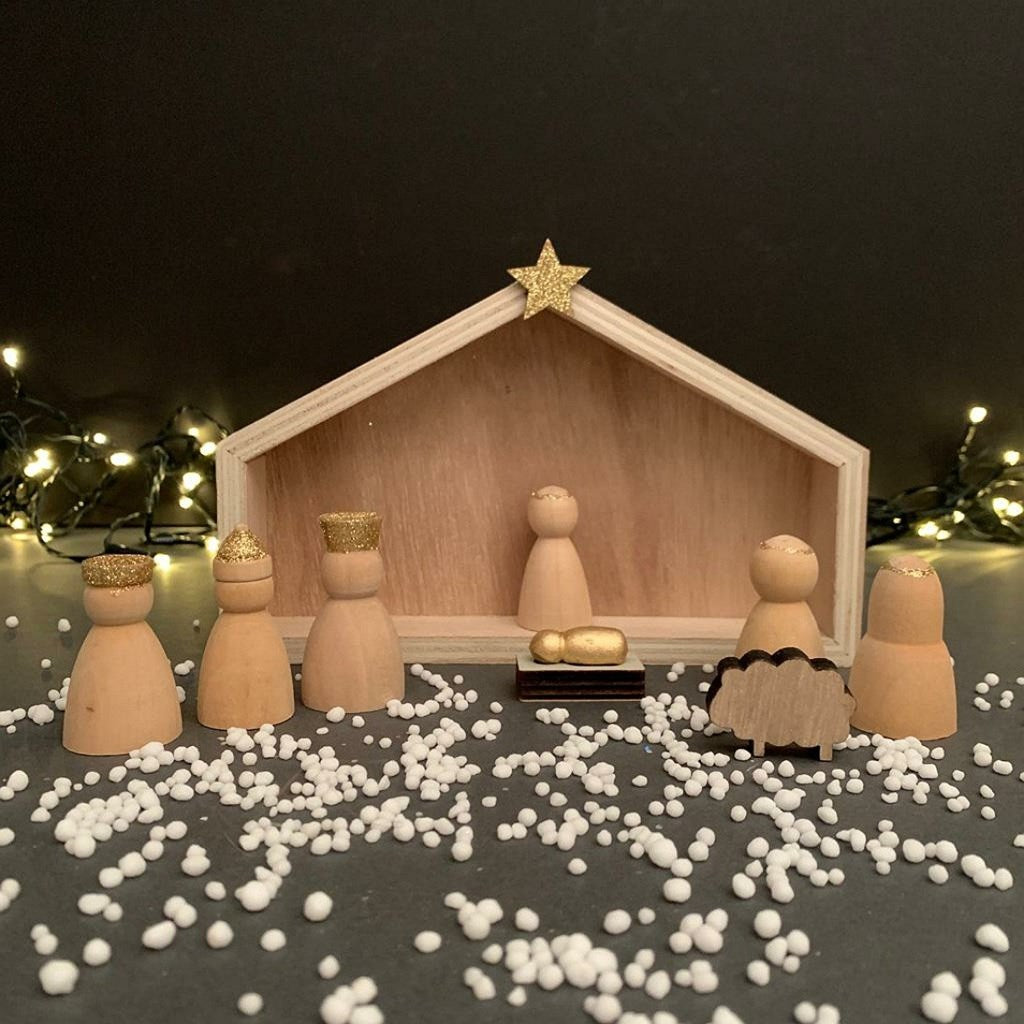A wooden nativity scene display with unpainted wooden figures, a stable backdrop, a gold star on top arranged on a dark surface scattered with white snow decorations.