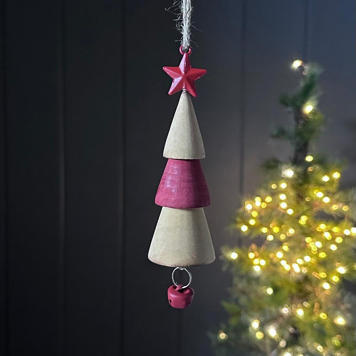 Decorative wooden Christmas tree ornament with a star on top and bell at the bottom, hanging against a dark background with a blurred Christmas tree in the background.