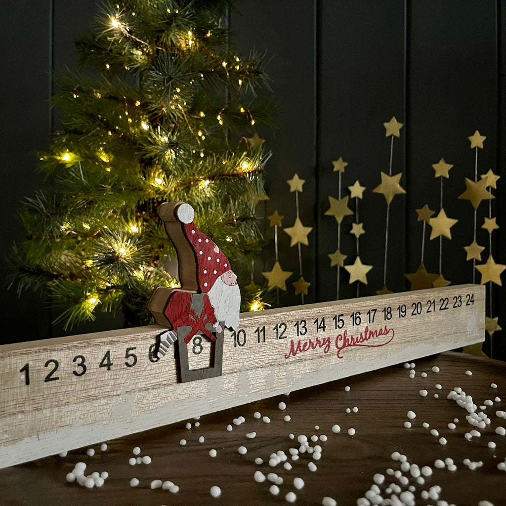 A Christmas advent countdown board with a wooden gnome figure sliding down a numbered track, styled on a tabletop with a lit Christmas tree and gold star decorations in the background.