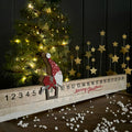 A Christmas advent countdown board with a wooden gnome figure sliding down a numbered track, styled on a tabletop with a lit Christmas tree and gold star decorations in the background.