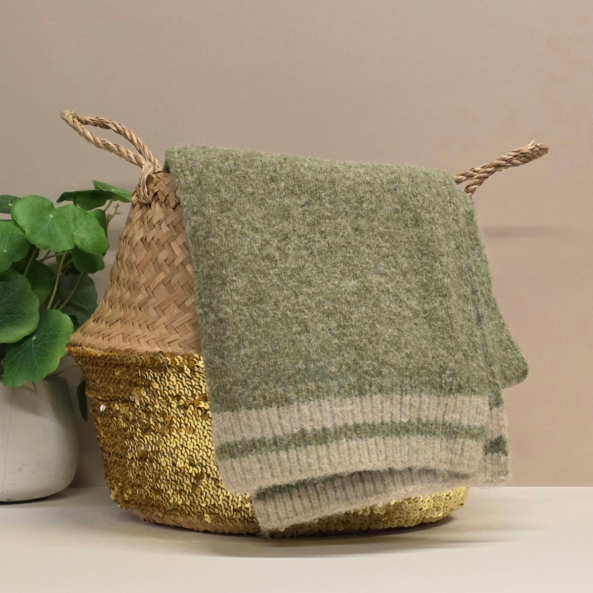 Decorative basket with a green knitted scarf draped over it on a neutral background. The scarf has a beige detail both ends. 