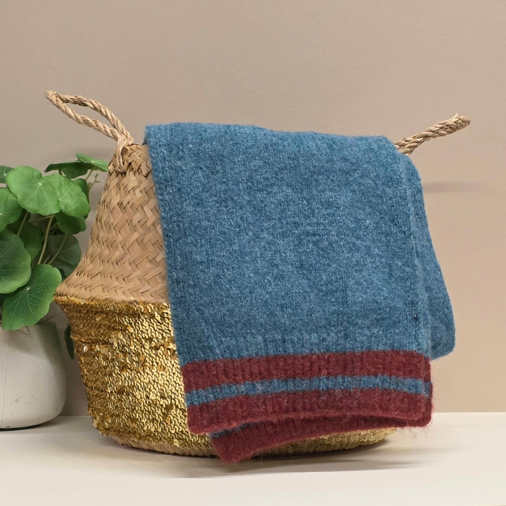 Blue knitted blanket scarf with red stripe draped over a woven basket on a neutral background
