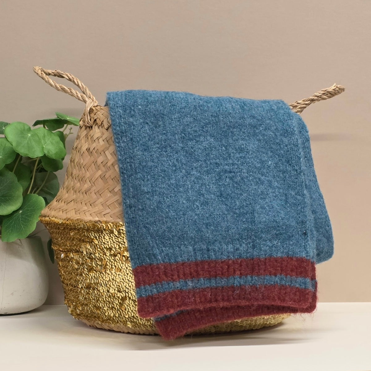 Blue knitted blanket scarf with red stripe draped over a woven basket on a neutral background