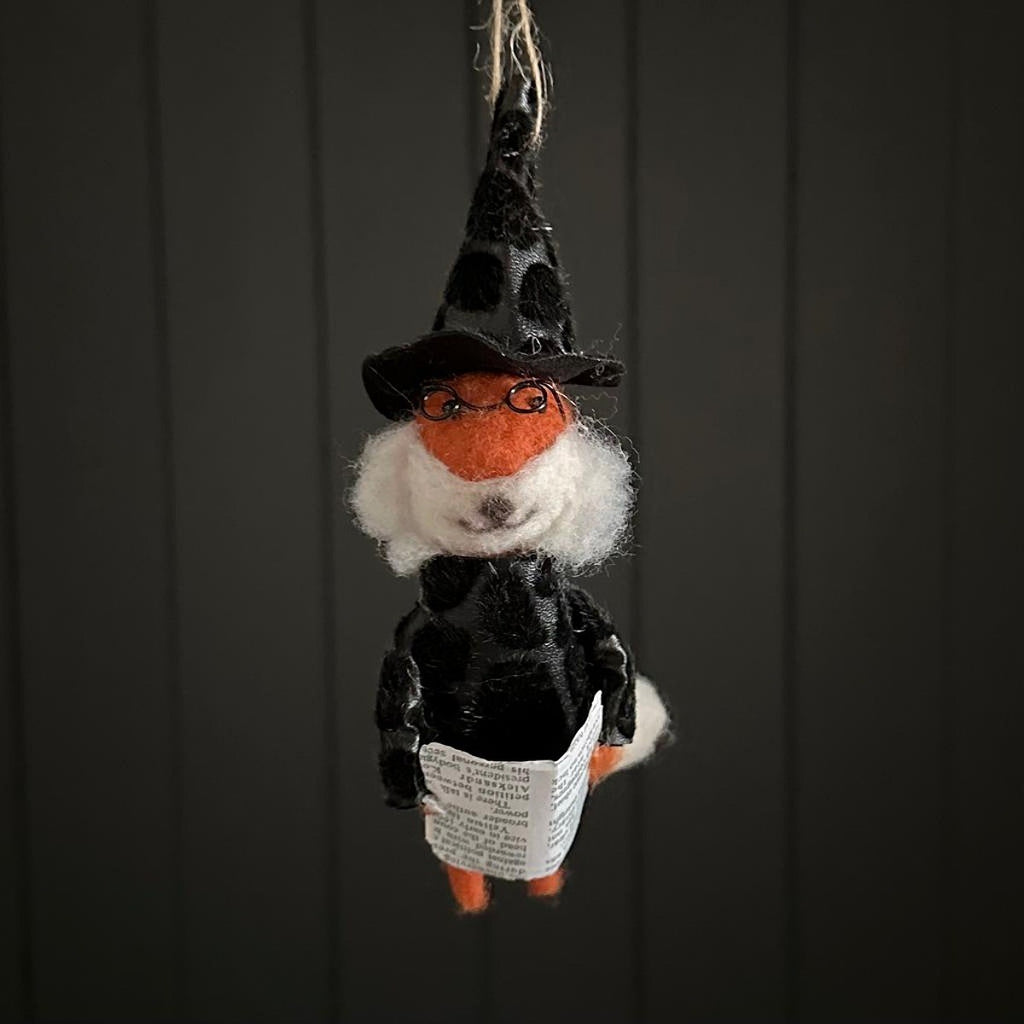 Hanging fox decoration dressed as a witch holding a magazine with a black hat and glasses on a dark background