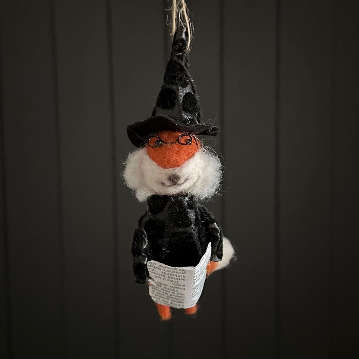 Hanging fox decoration dressed as a witch holding a magazine with a black hat and glasses on a dark background
