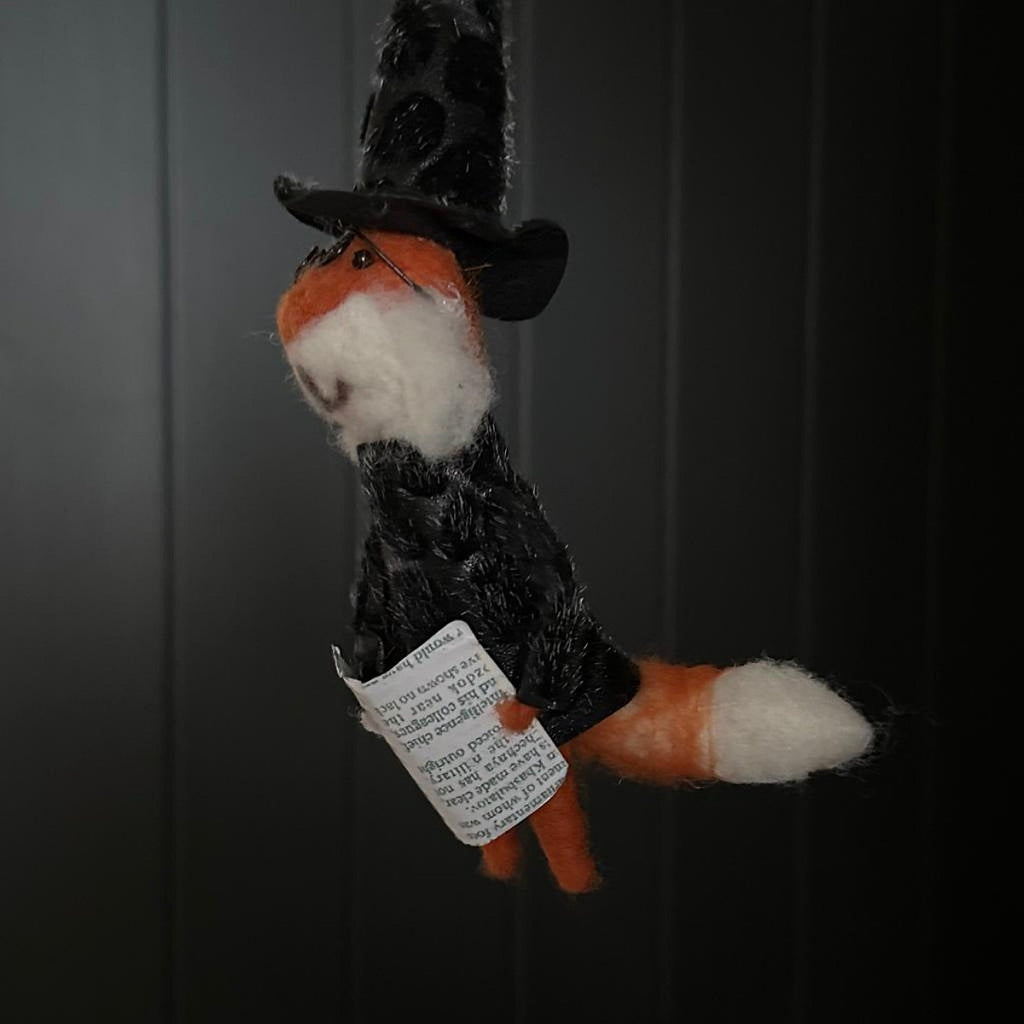 Hanging fox decoration dressed as a witch holding a magazine with a black hat and glasses on a dark background
