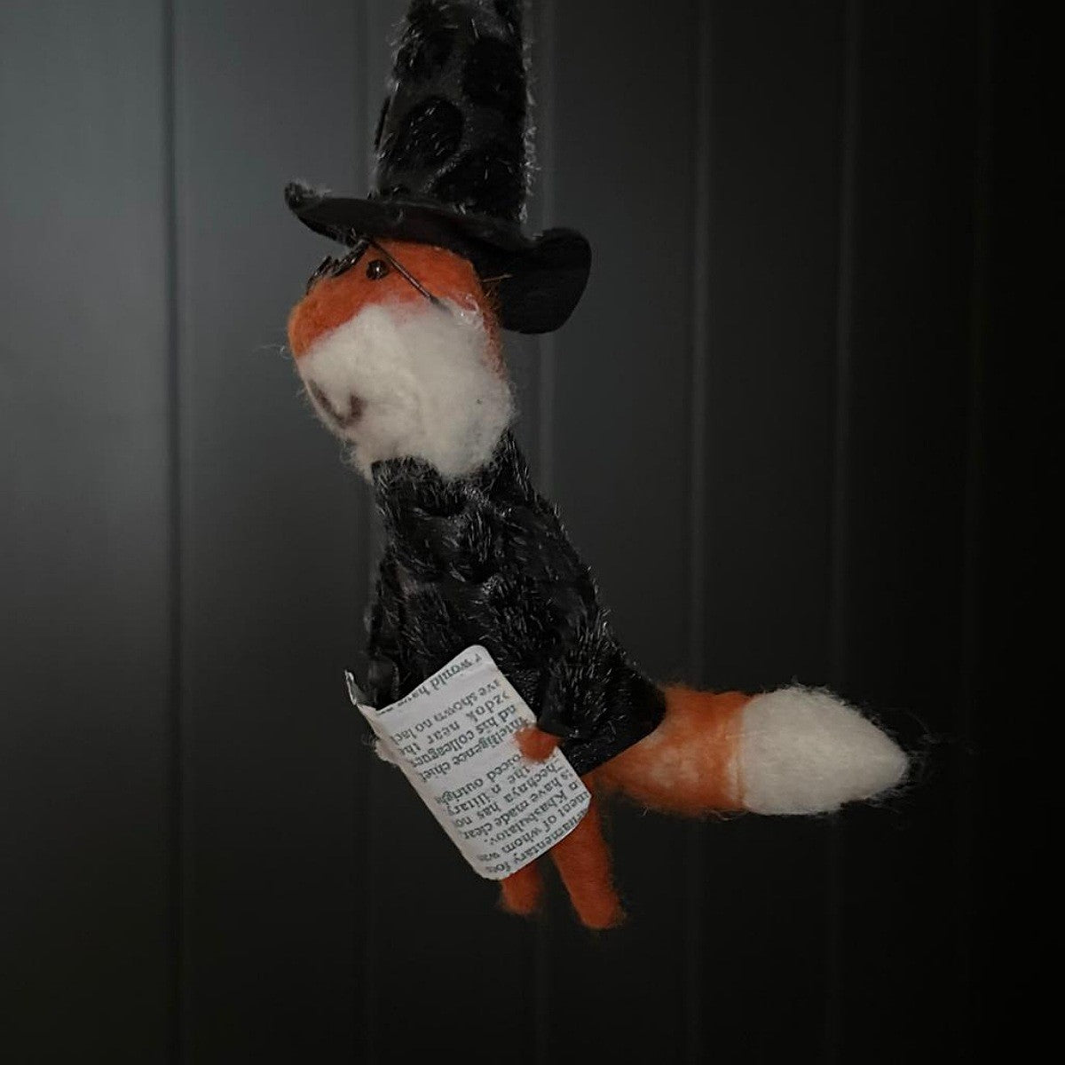 Hanging fox decoration dressed as a witch holding a magazine with a black hat and glasses on a dark background