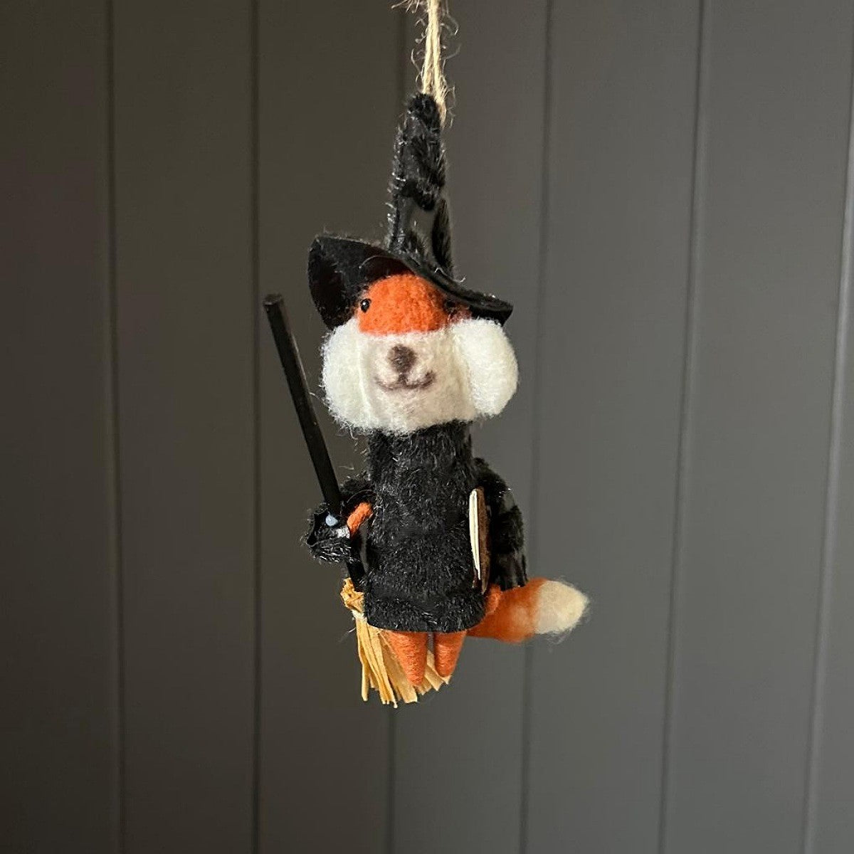 A small felted wool witch fox hanging ornament, featuring an orange and white dog body dressed in a black witch hat and cloak, holding a broom, set against a dark grey panelled background.