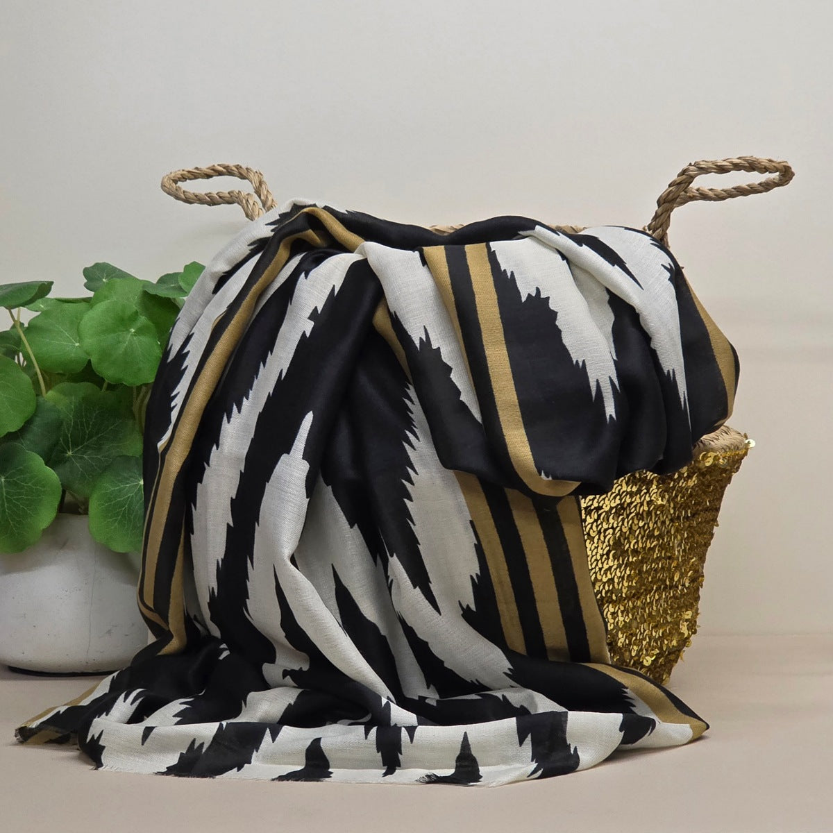 A black, white, and gold patterned scarf draped over a woven gold basket with rope handles, next to a potted green plant against a neutral beige background.