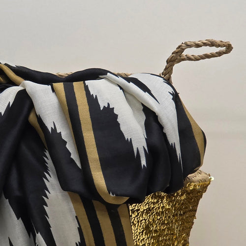 A draped scarf with a bold black, white, and gold ikat-inspired pattern, styled over a woven basket against a neutral background.