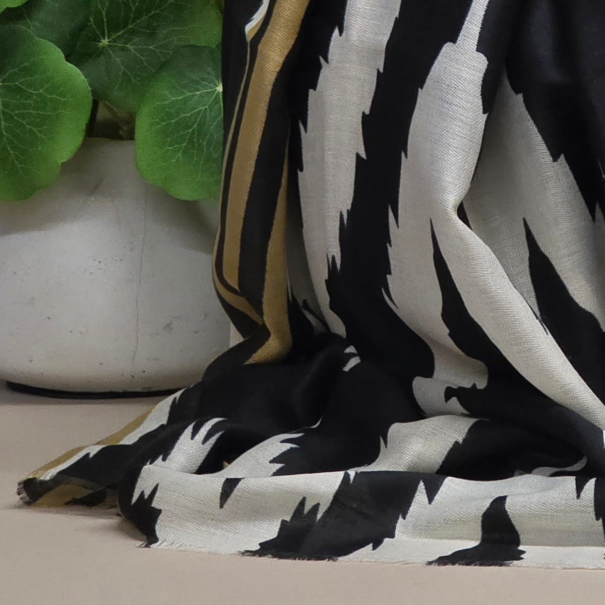 A draped black and white patterned scarf with a gold border, styled next to a white ceramic pot with green leaves.
