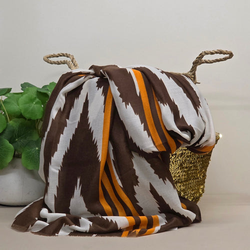 A zig zag patterned throw scarf draped over a woven basket with rope handles, featuring a brown and white ikat-style design with orange stripes, set against a neutral background with a potted plant.