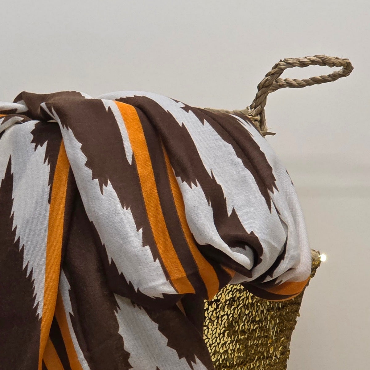 A draped patterned scarf with brown and white zigzag stripes and orange vertical bands, shown on a mannequin stand with a rope handle against a plain light background.