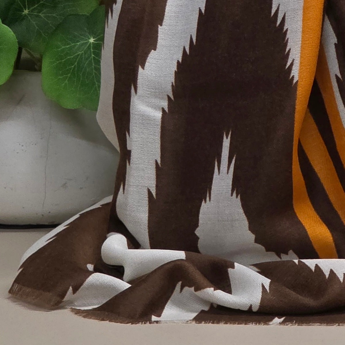A draped scarf with a brown and white ikat-style geometric pattern and a vertical orange stripe, styled next to a white planter with green leaves.