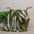 A green and white ikat patterned scarf draped over a woven basket with a gold sequin trim, next to a green potted plant against a neutral beige background.
