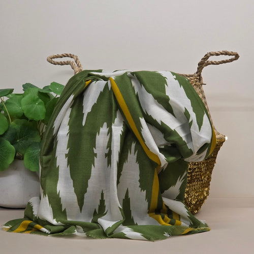 A green and white ikat patterned scarf draped over a woven basket with a gold sequin trim, next to a green potted plant against a neutral beige background.